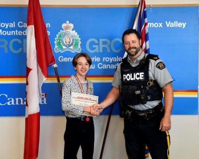 Rylan and Sgt. Rob Brennan standing in front of a sign for the Comox Valley RCMP and Canada flag. Sgt. Brennan is presenting Rylan with a certificate and they are shaking hands.